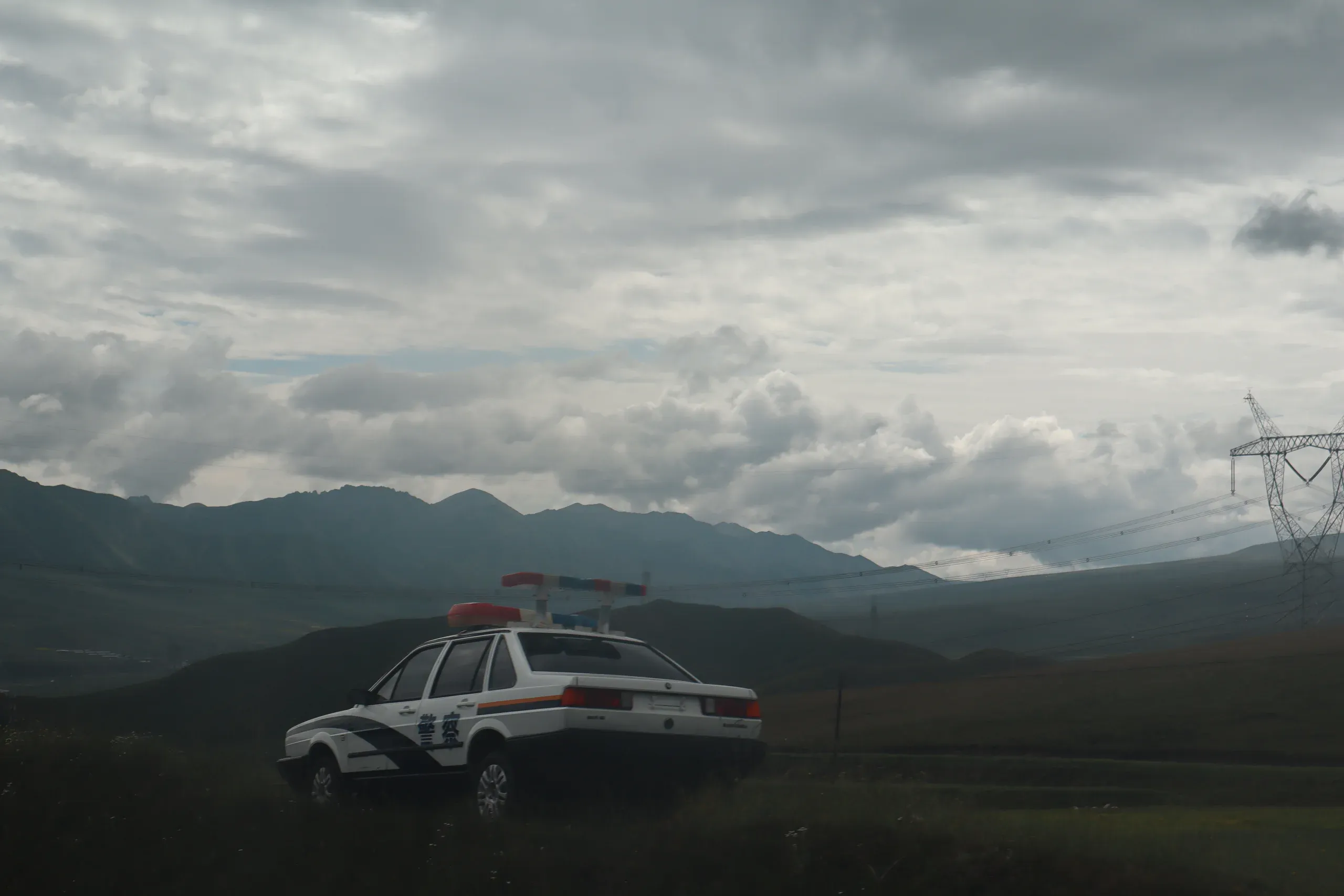 乌云下的高原公路旁停着一辆警车，远处是山脉和输电塔